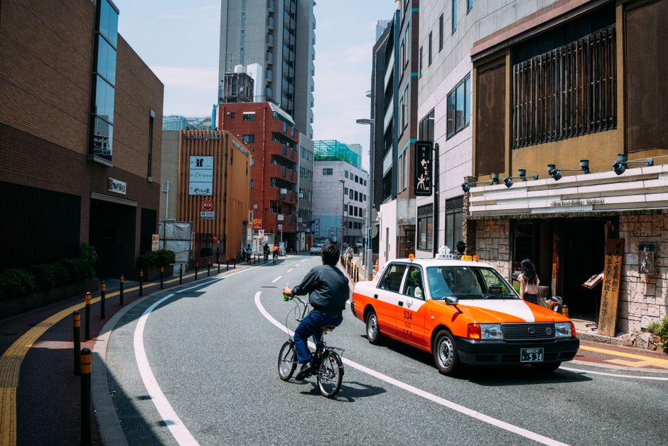 Mobilidade Urbana — Cyclist navigates city street alongside taxi on a sunny day