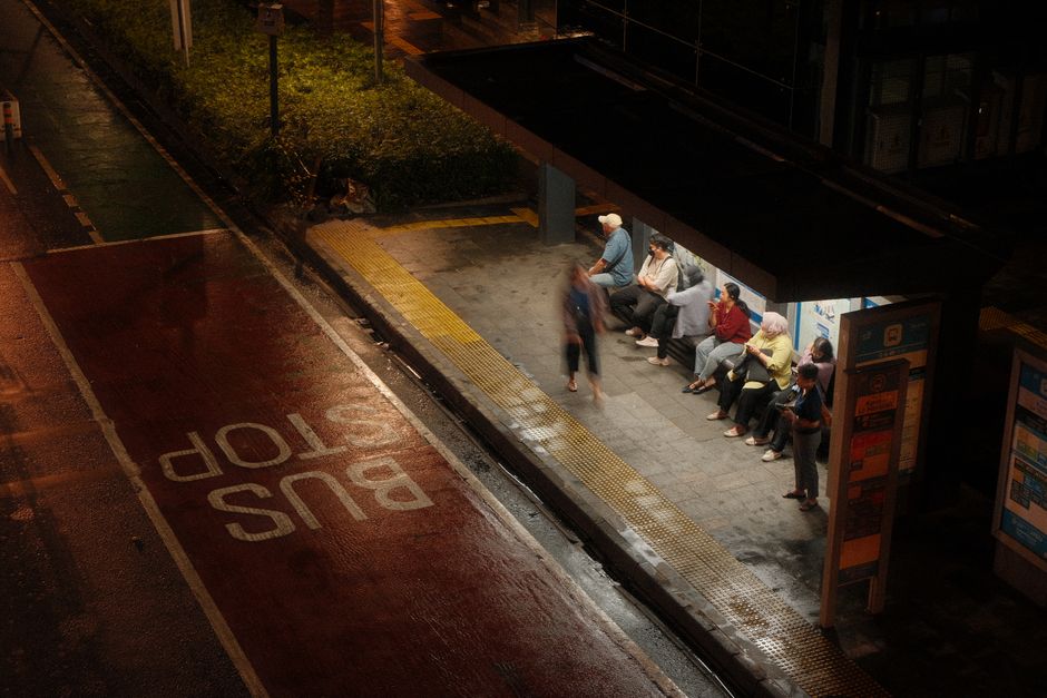 Mobilidade Urbana — A dimly lit bus stop at night with diverse passengers waiting under shelter.