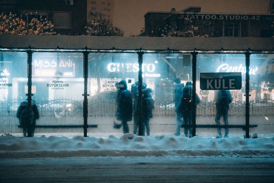 Mobilidade Urbana — People waiting at a snowy bus stop in Konya, Türkiye under warm night lights.