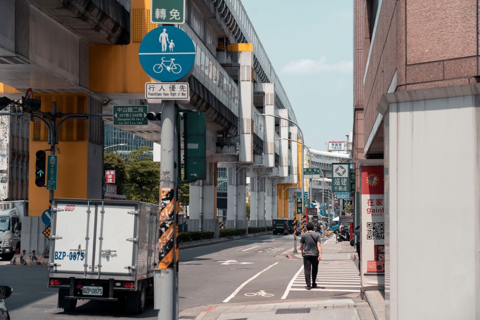 Mobilidade Urbana — Bustling street in New Taipei City under elevated train tracks with signs and pedestrians.